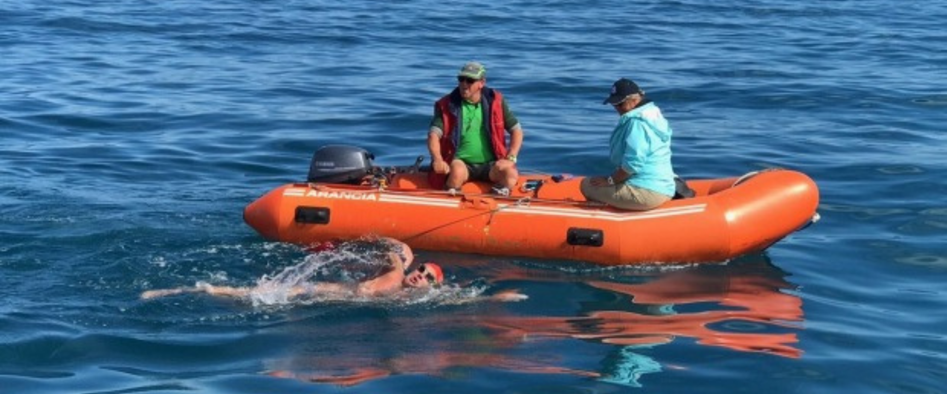 Marty swimming in the water in a red swimming cap and Zoggs Predator Goggles. Swimming next to a orange support boat with a man and woman seated in the boat